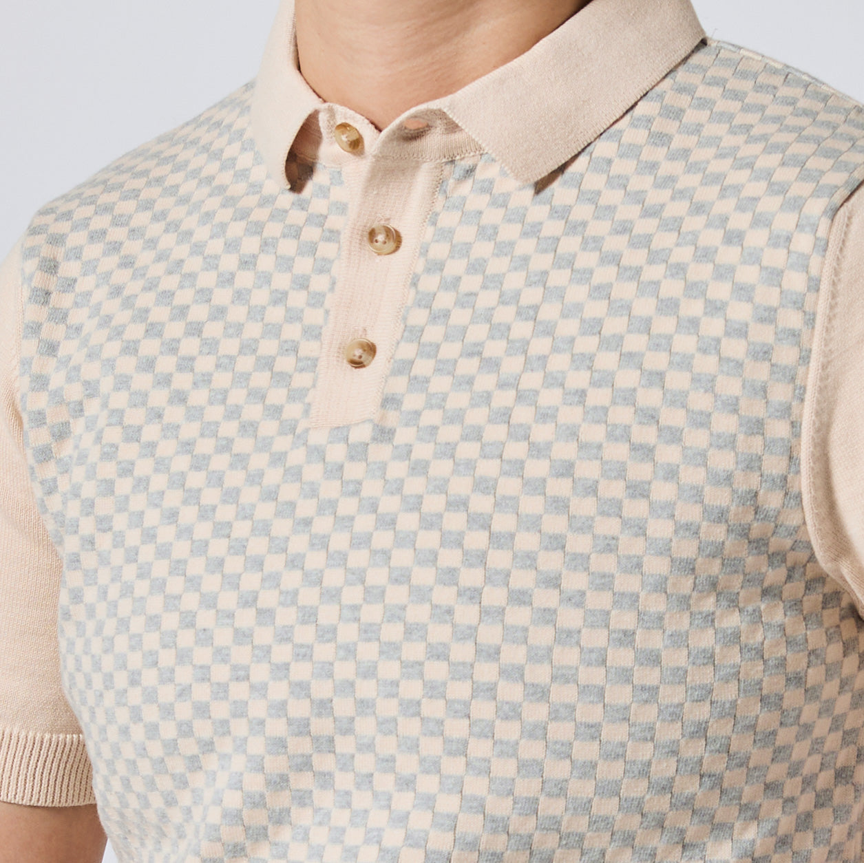 Close-up of a person wearing a short-sleeve knit polo shirt with a beige and light blue checkerboard pattern, a solid beige collar, and three gold buttons. Color: Saltstone Checker