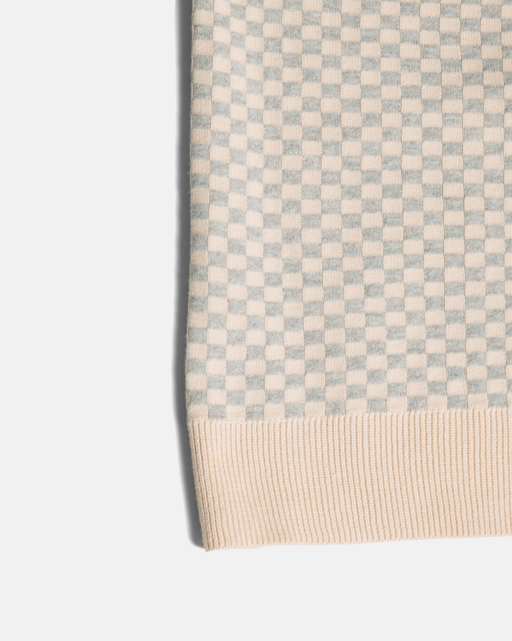 Close-up of a ribbed knit fabric with a small checkerboard pattern in light gray and cream, finished with a cream ribbed hem. Color: Saltstone Checker