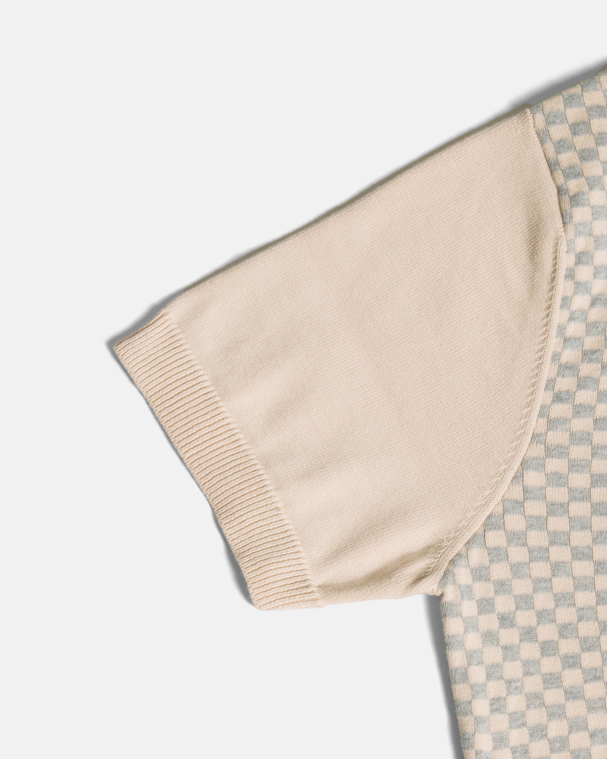 Close-up of a short-sleeve knit top showing a solid beige ribbed sleeve cuff and part of a beige and light gray checkerboard patterned body, on a light gray background. Color: Saltstone Checker