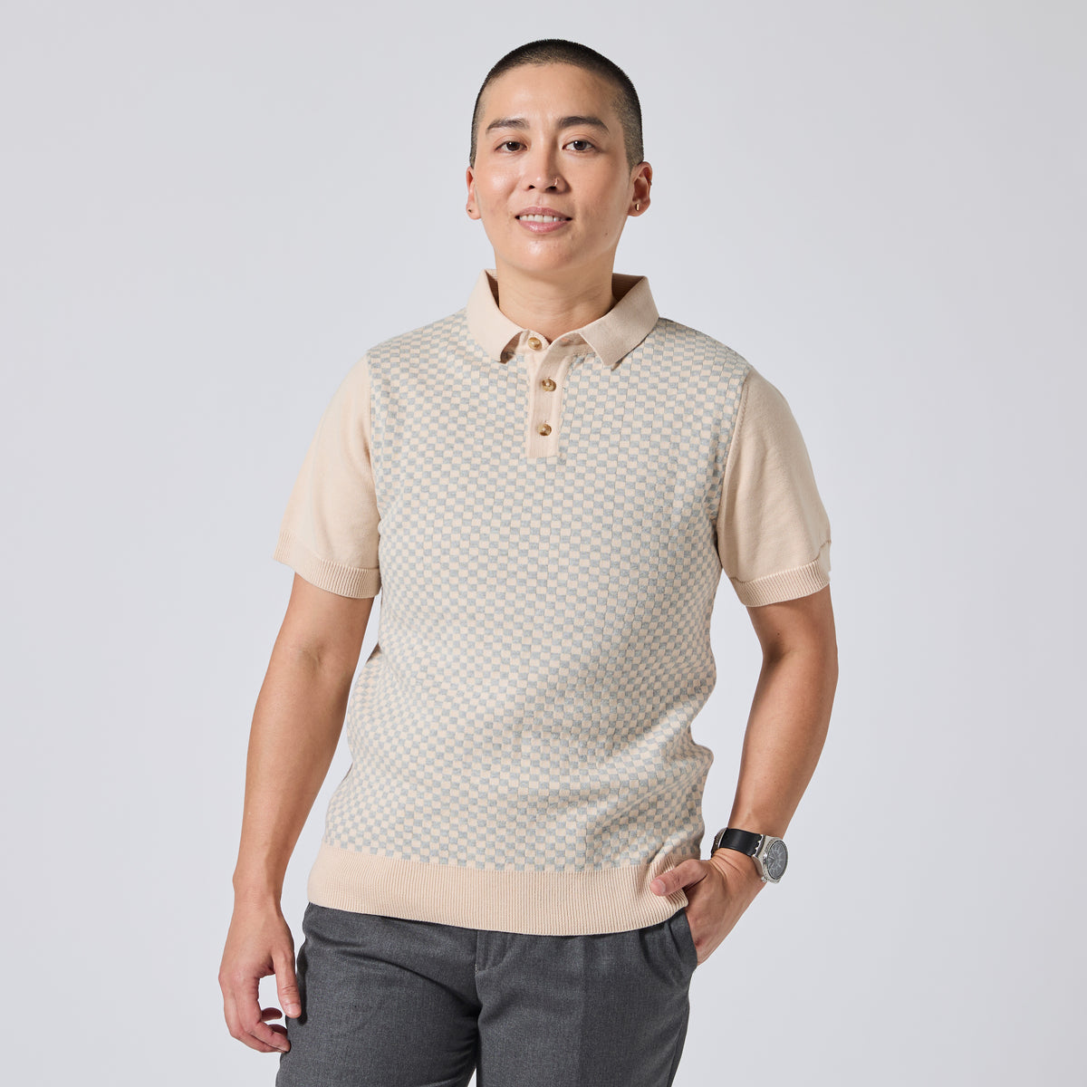 Model wearing a beige and off-white patterned knit polo vest over a matching beige polo shirt, paired with dark gray trousers, against a light gray studio background. Color: Saltstone Checker