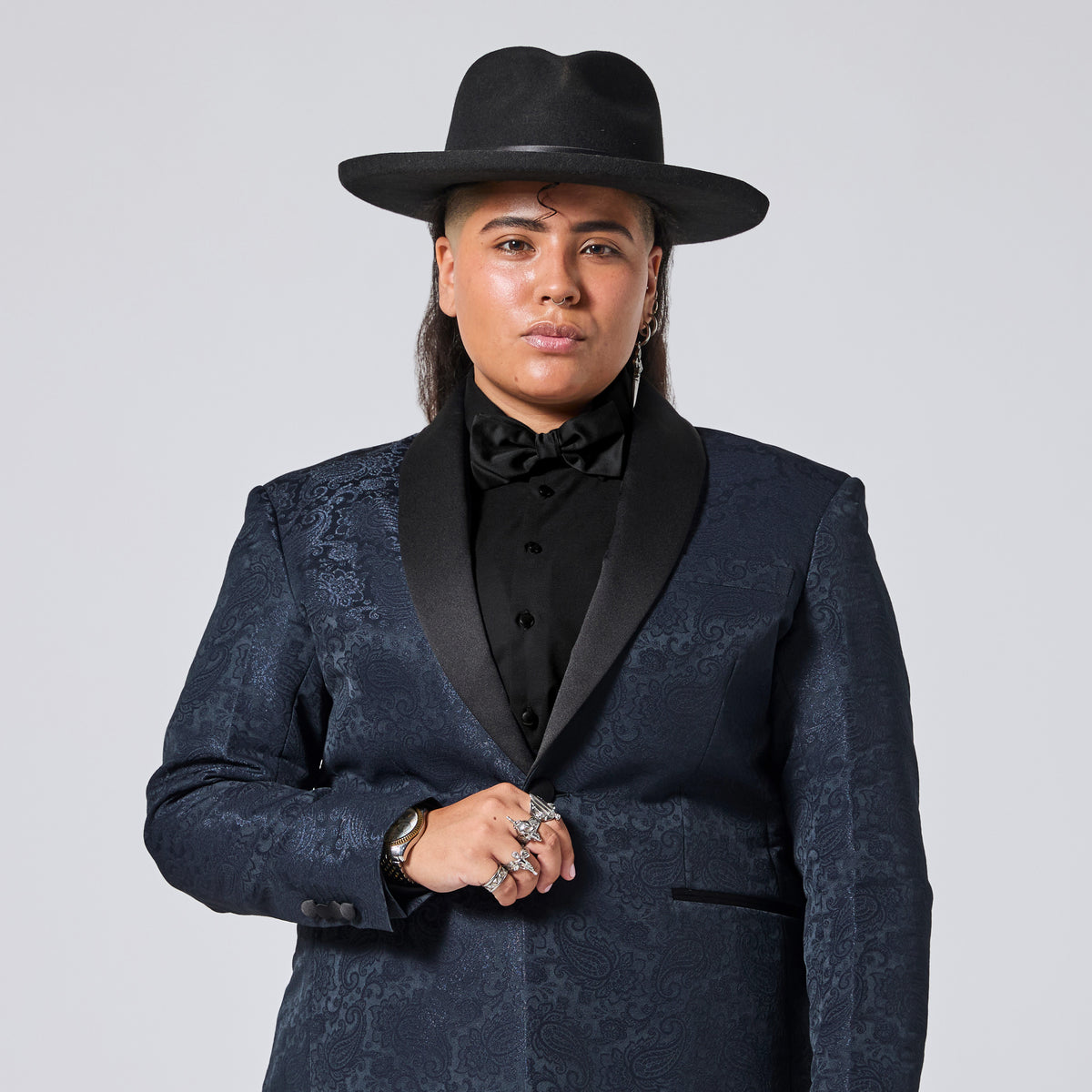 A person wearing a dark navy patterned tuxedo jacket with black satin lapels, a black shirt, a black bow tie, a black wide-brim hat, and multiple rings, photographed against a light gray studio background. Color: Sapphire Brocade
