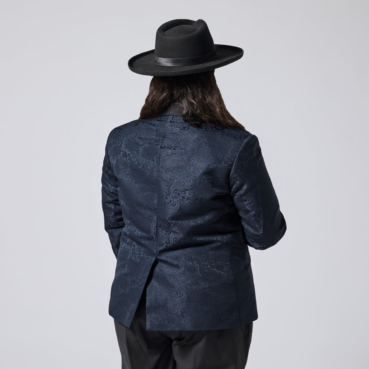 A person shown from the back wearing a dark navy patterned blazer, black pants, and a black wide-brim hat against a light gray studio background. Color: Sapphire Brocade