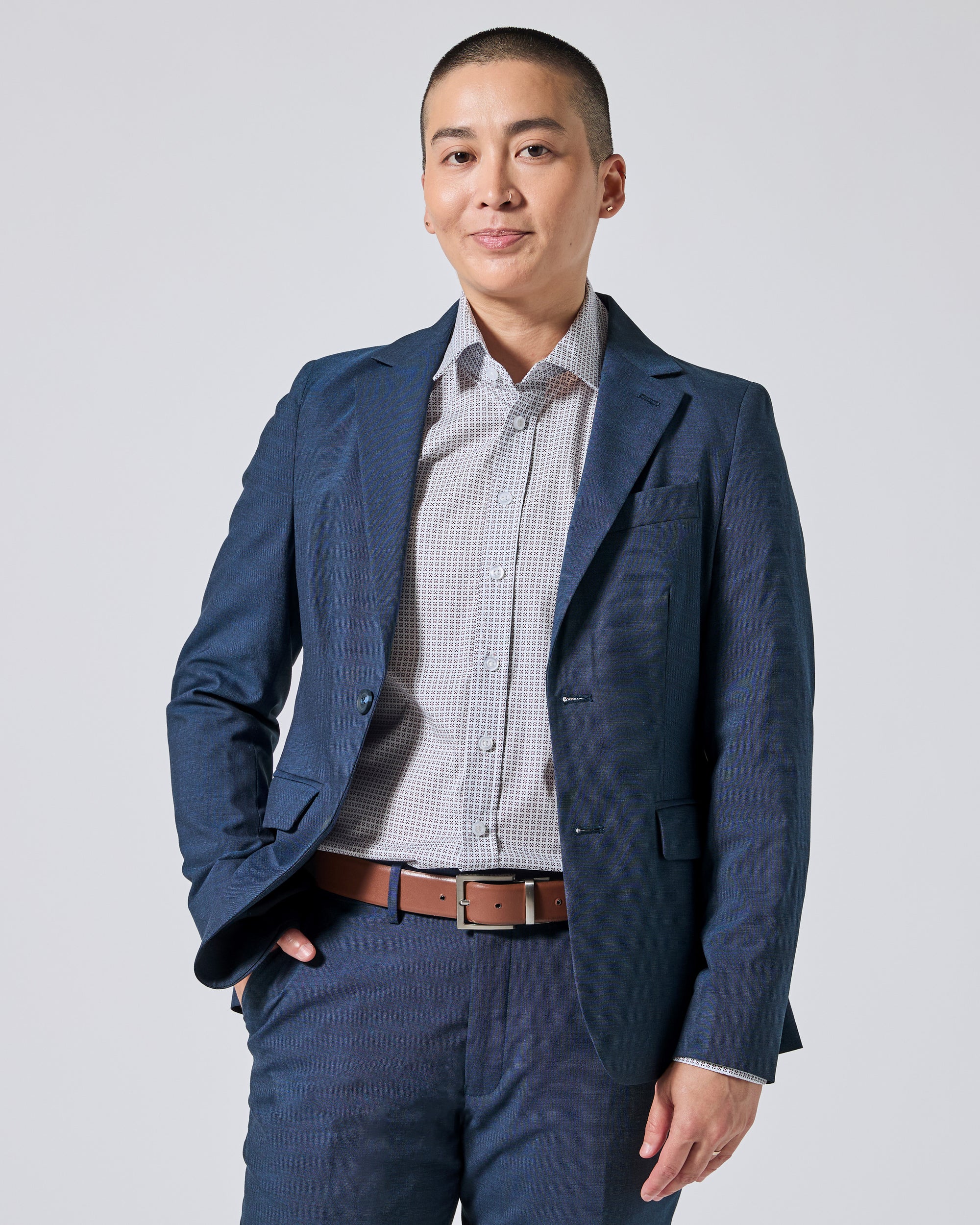 Person wearing an Atlantic blue blazer and matching trousers, paired with a light gray checkered button-down shirt and a brown leather belt, standing against a light gray studio background. Color: Atlantic Blue