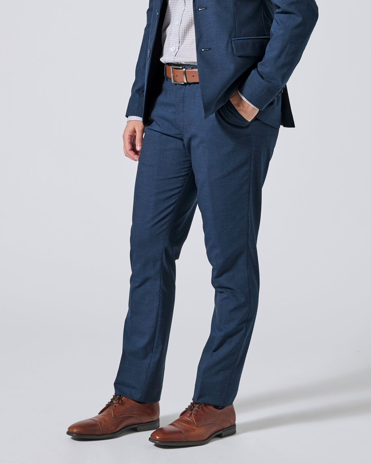 Person wearing Atlantic blue trousers, a matching navy blazer, a white shirt, a brown belt, and brown leather dress shoes, shown from the waist down on a light gray studio background. Color: Atlantic Blue