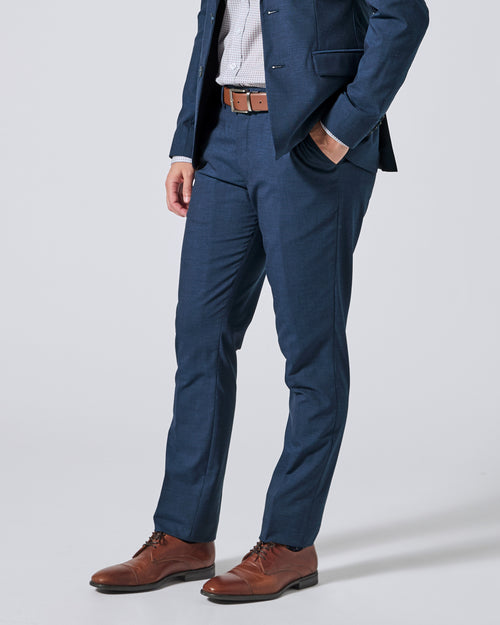 Person wearing Atlantic blue trousers, a matching navy blazer, a white shirt, a brown belt, and brown leather dress shoes, shown from the waist down on a light gray studio background. Color: Atlantic Blue