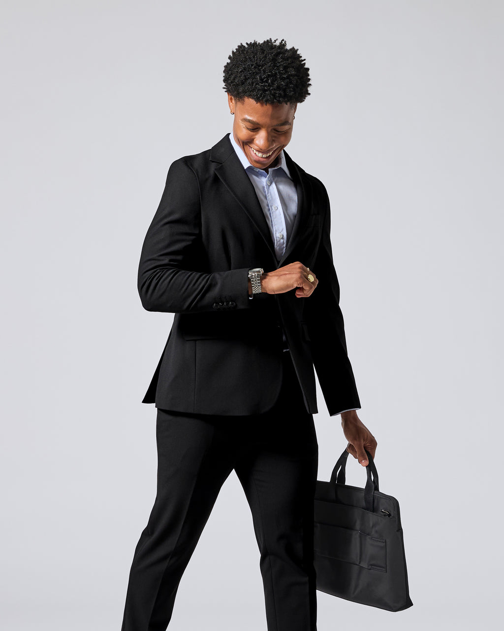 Person wearing a black two-piece suit, light blue dress shirt, and a gold ring, holding a black briefcase against a solid gray background. Color: Obsidian