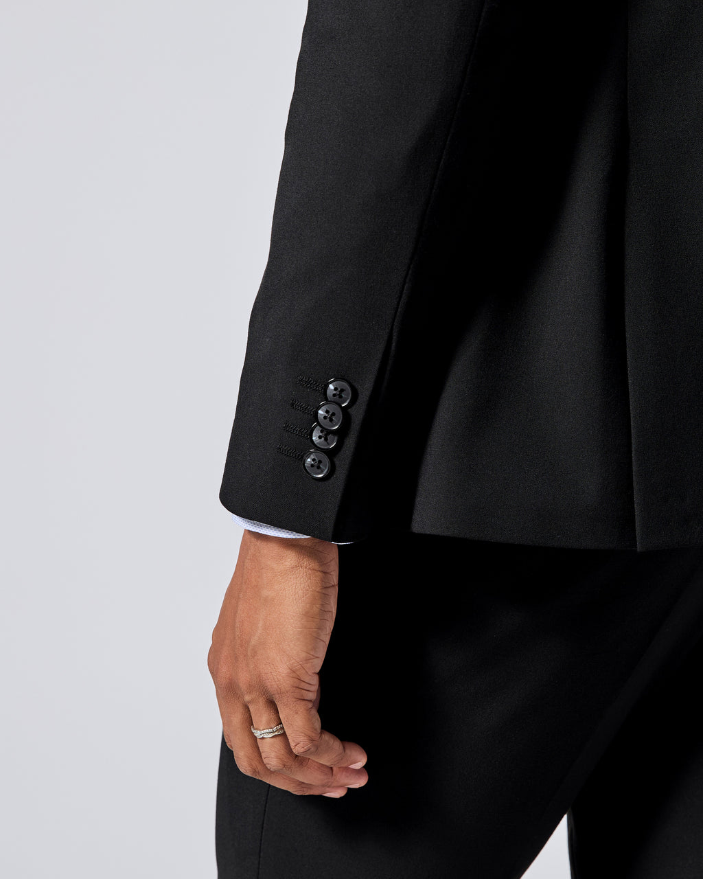 Close-up of a black tailored jacket sleeve with four dark buttons, worn with matching black trousers and a white shirt cuff visible at the cufflink area, against a light gray studio background. Color: Obsidian