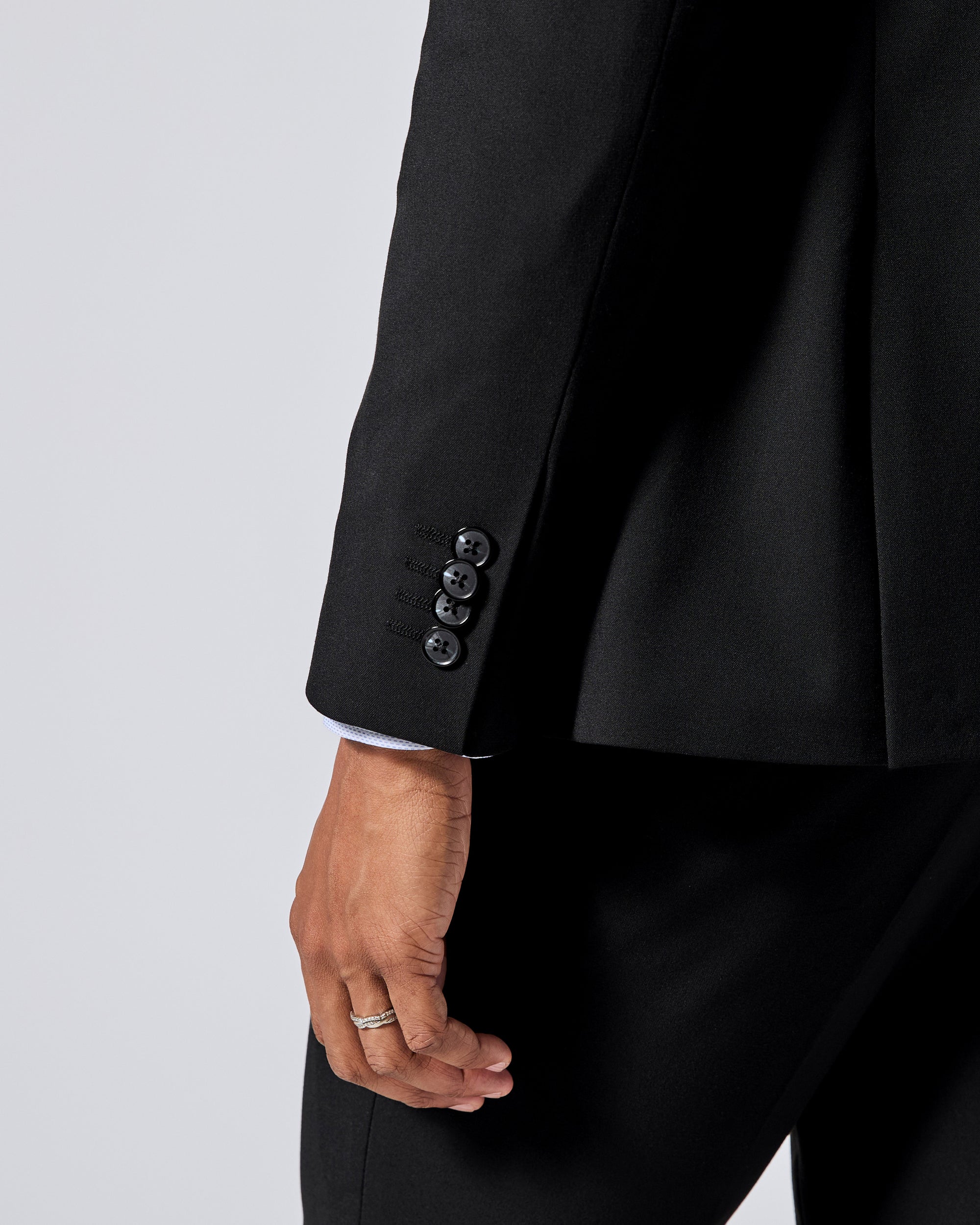 Close-up of a black tailored jacket sleeve with four dark buttons, worn with matching black trousers and a white shirt cuff visible at the cufflink area, against a light gray studio background. Color: Obsidian