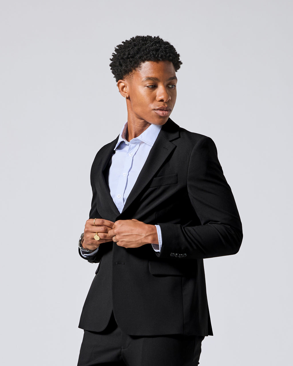 Person wearing a black tailored blazer over a light blue dress shirt, standing against a plain light gray studio background and buttoning his blazer. Color: Obsidian