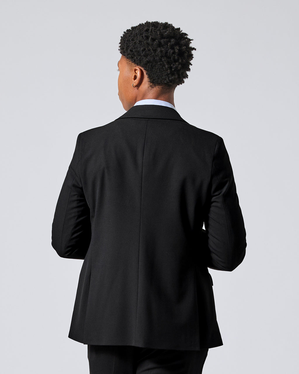 Person viewed from the back wearing a black suit jacket over a white collared shirt, standing against a light gray studio background. Color: Obsidian
