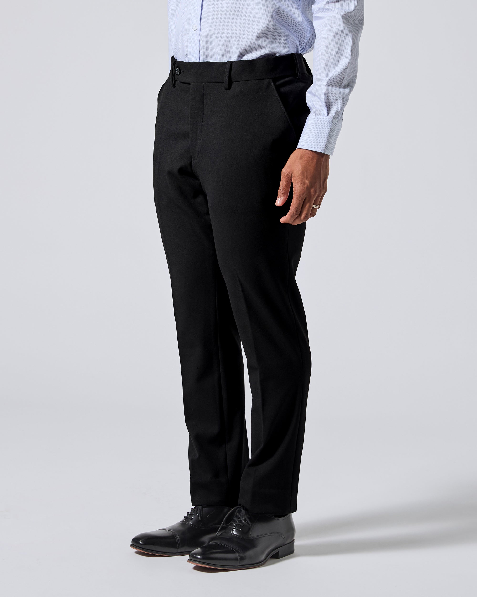A person wearing black dress pants with a white dress shirt and black dress shoes, shown from the waist down on a light gray studio background. Color: Obsidian