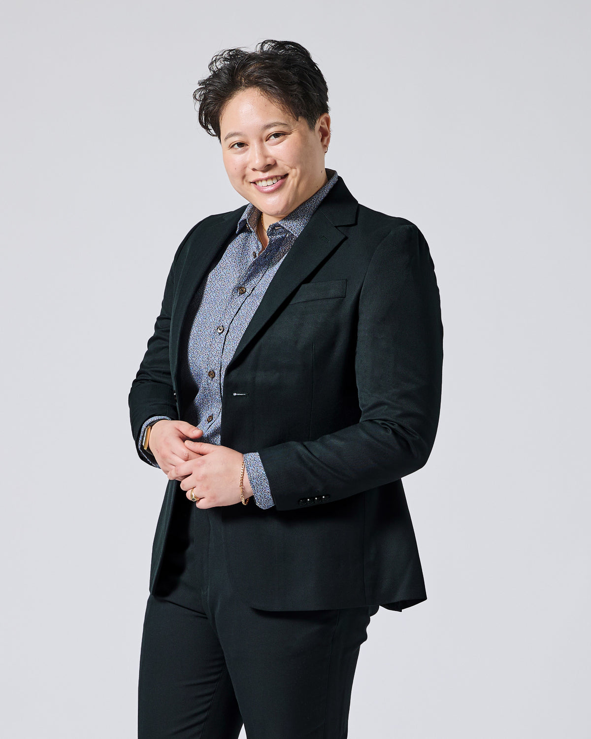 Person wearing a Phantom Green Twill Scout blazer over a blue patterned button-down shirt, paired with black trousers, standing against a solid light gray background. Color: Phantom Green Twill