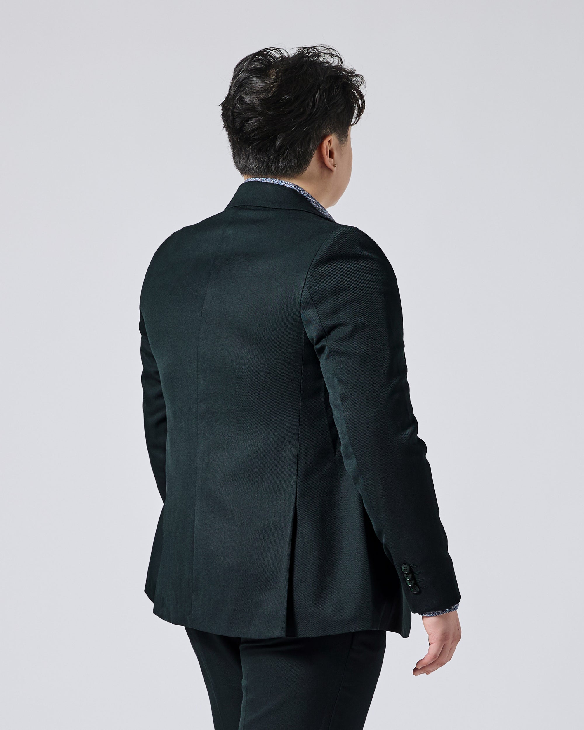 Person viewed from behind wearing a Phantom Green Twill blazer over a light blue collared shirt, set against a plain light gray studio background. Color: Phantom Green Twill