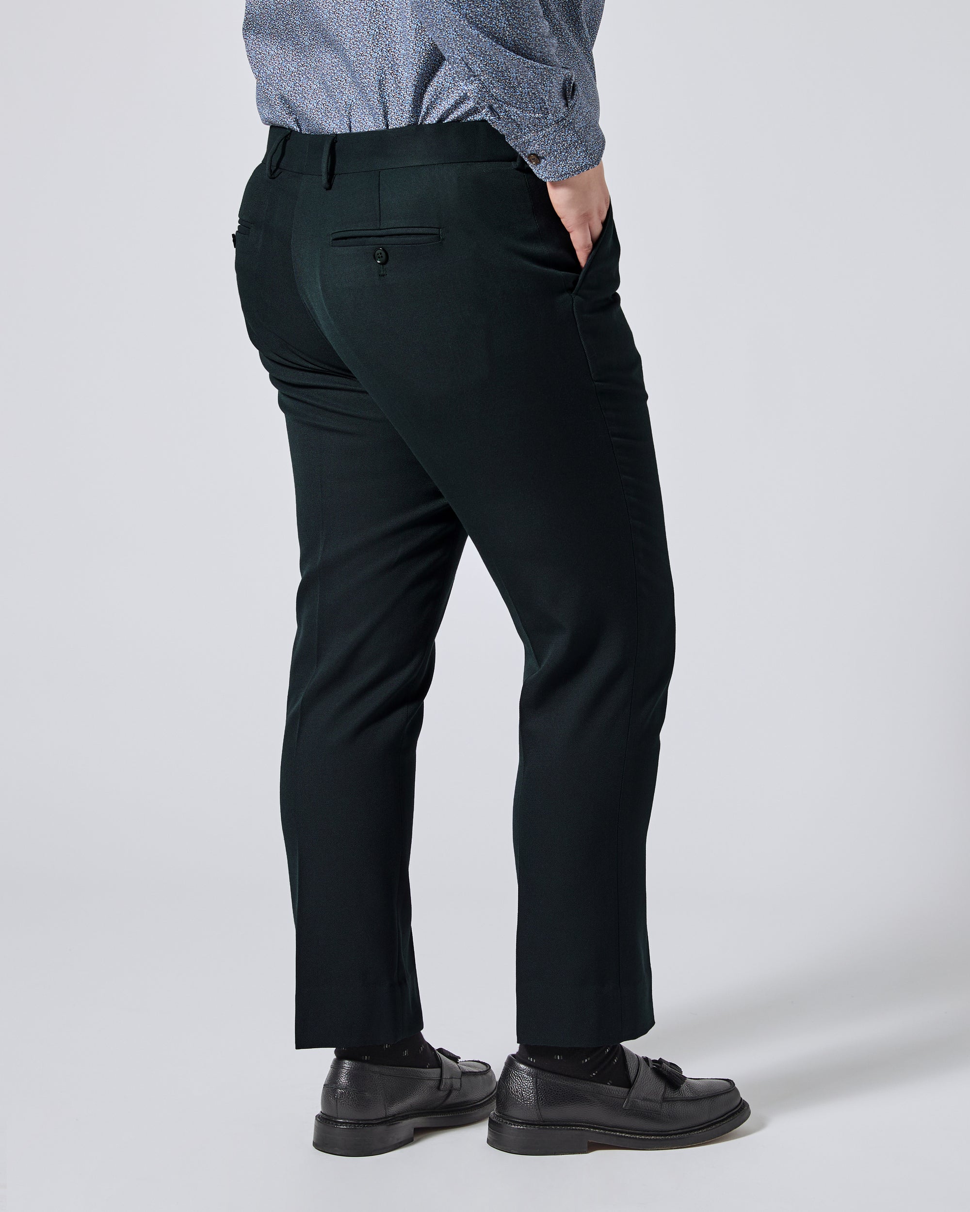 A person shown from the back wearing dark green trousers, a blue patterned shirt, and black loafers against a light gray studio background. Color: Phantom Green Twill