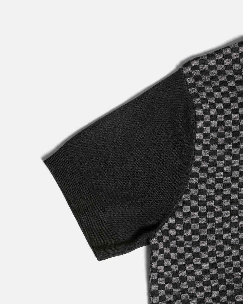 Close-up of a short-sleeve knit top showing a solid black sleeve and a black-and-gray checkerboard patterned body, photographed on a light gray background. Color: Shadow Checker
