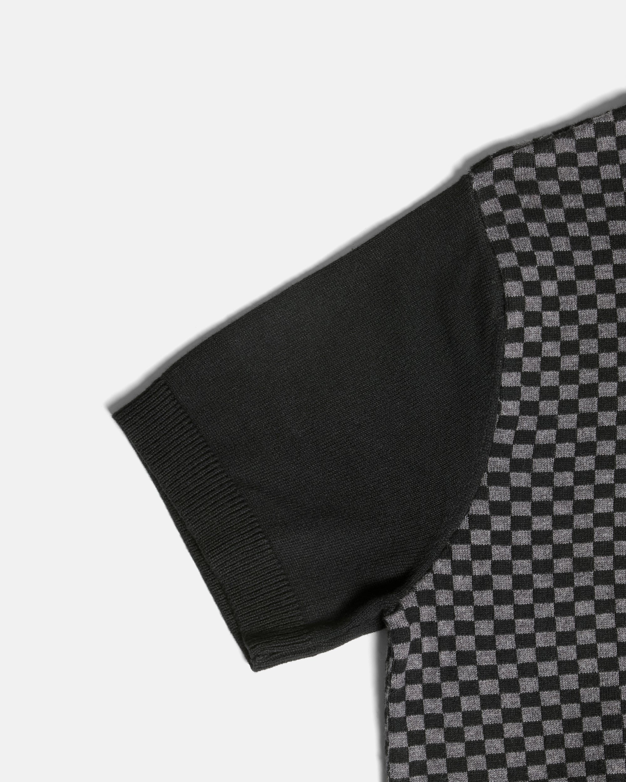 Close-up of a short-sleeve knit top showing a solid black sleeve and a black-and-gray checkerboard patterned body, photographed on a light gray background. Color: Shadow Checker