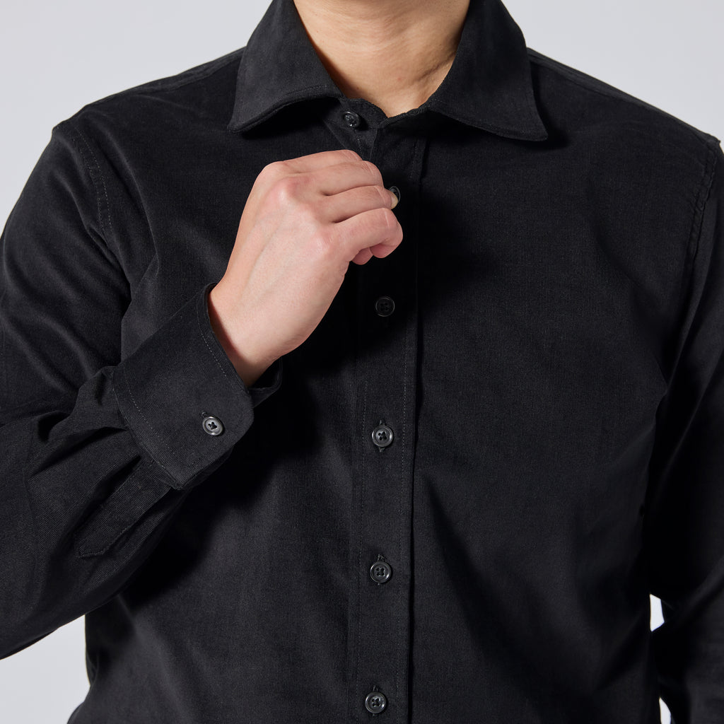A close-up of a person wearing a black long-sleeve button-up shirt, adjusting the collar and buttons with one hand against a light gray background. Color: Shadow Corduroy