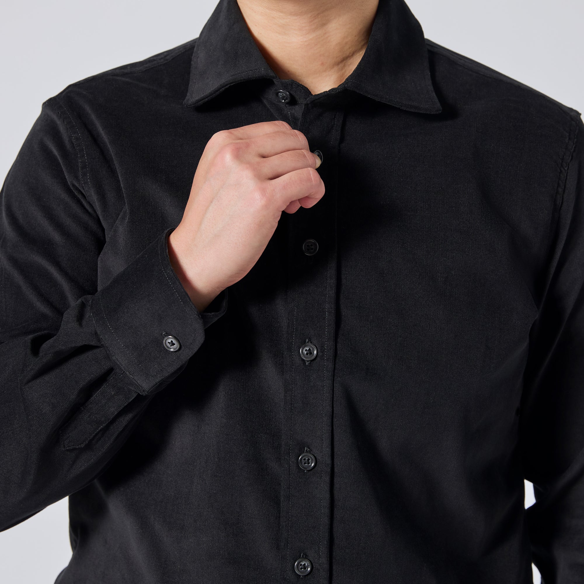 A close-up of a person wearing a black long-sleeve button-up shirt, adjusting the collar and buttons with one hand against a light gray background. Color: Shadow Corduroy