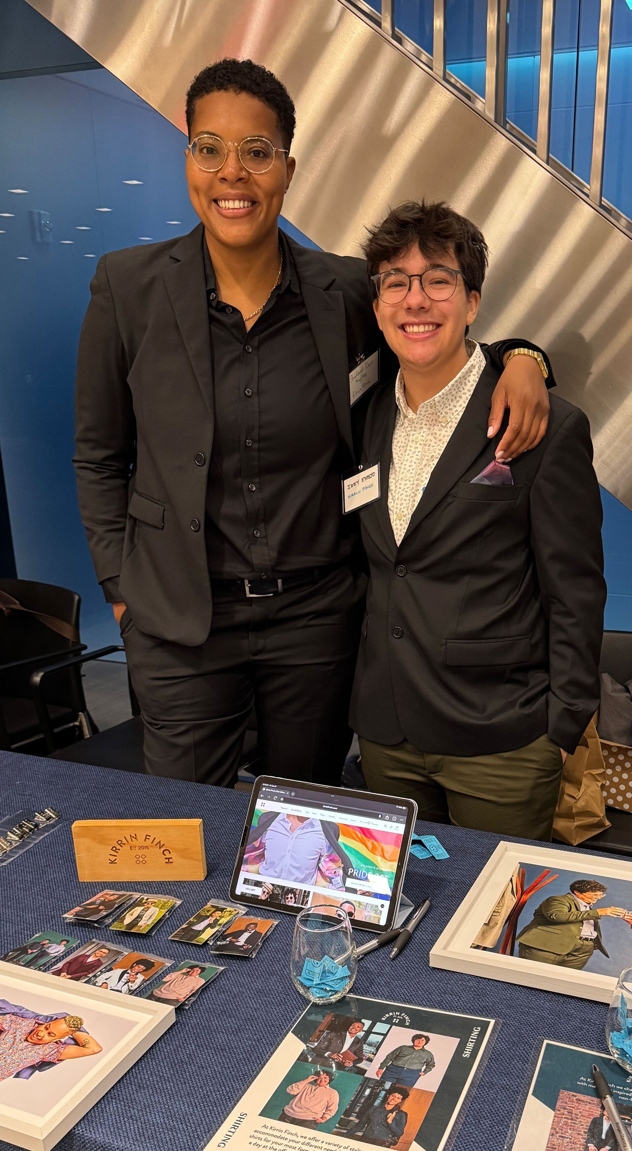Two people standing behind a table with art and a laptop, in front of a blue wall with a staircase.