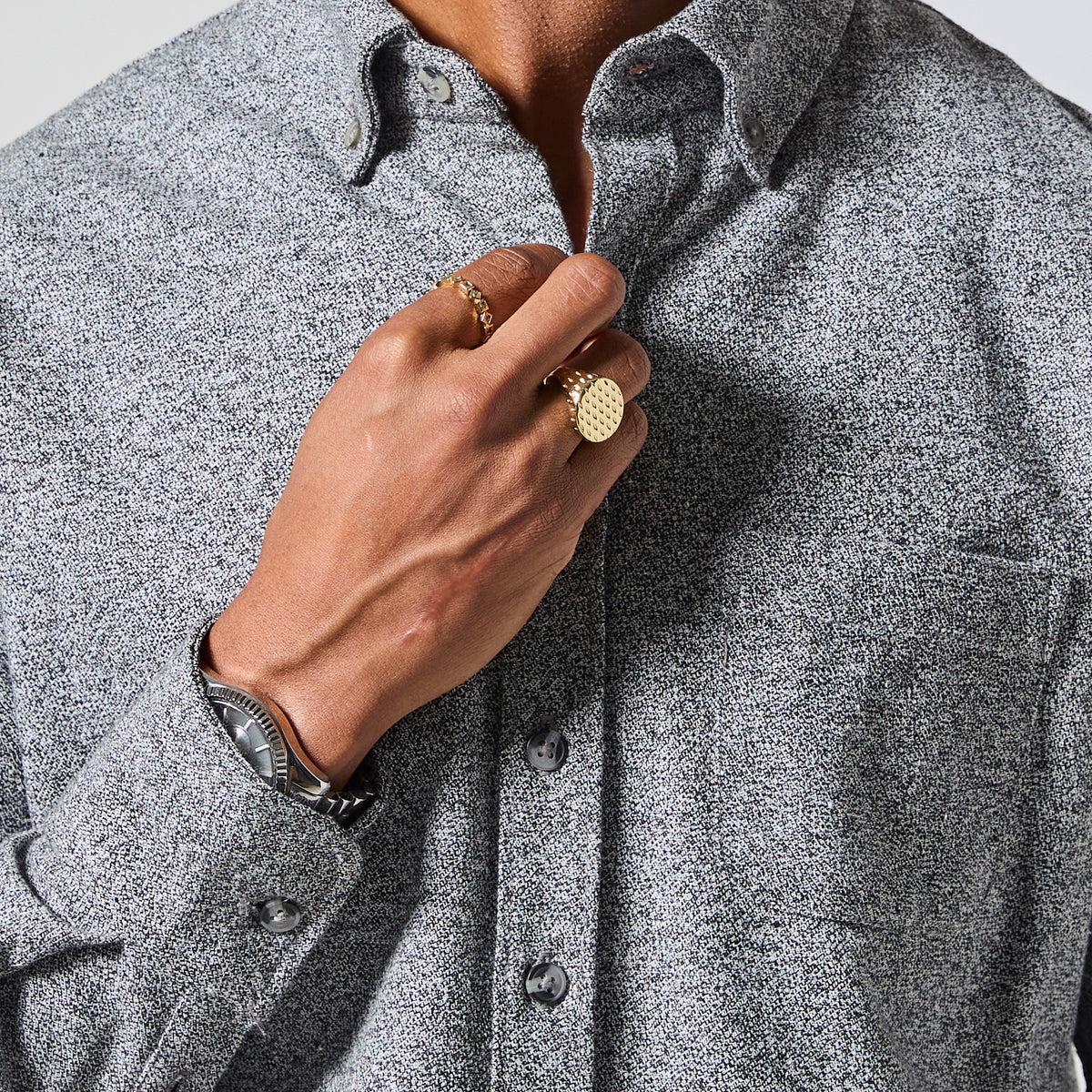 Close-up of a person wearing a textured gray long-sleeve button-down shirt, with one hand adjusting the collar and wearing gold rings and a watch. Color: Static
