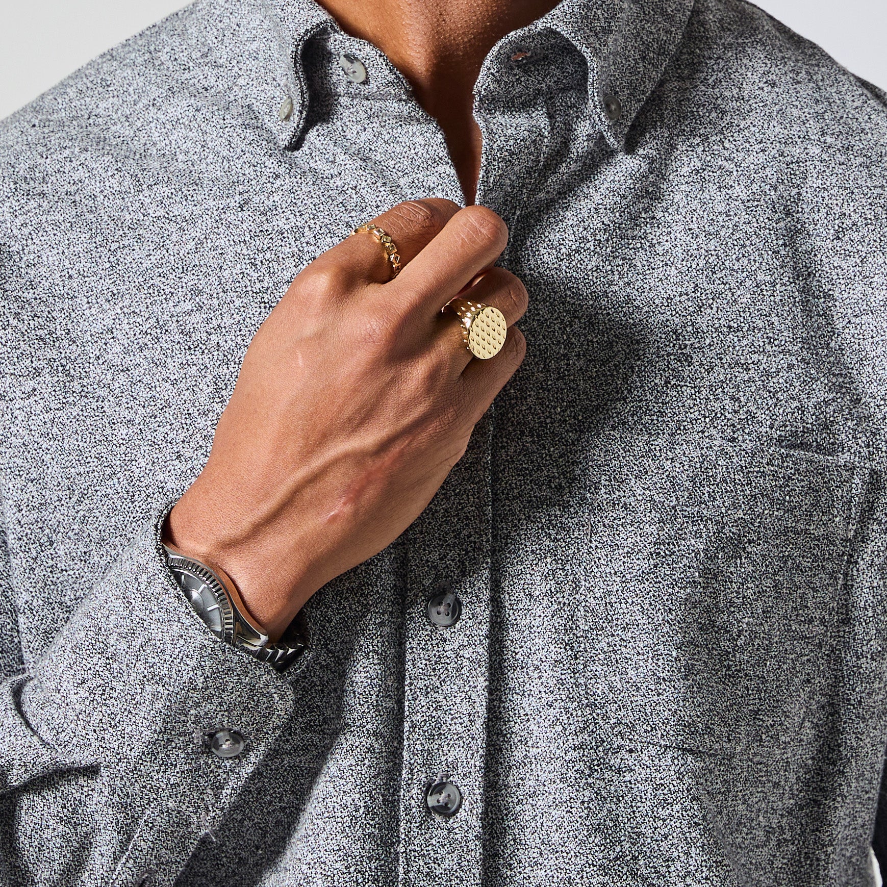 Close-up of a person wearing a textured gray long-sleeve button-down shirt, with one hand adjusting the collar and wearing gold rings and a watch. Color: Static