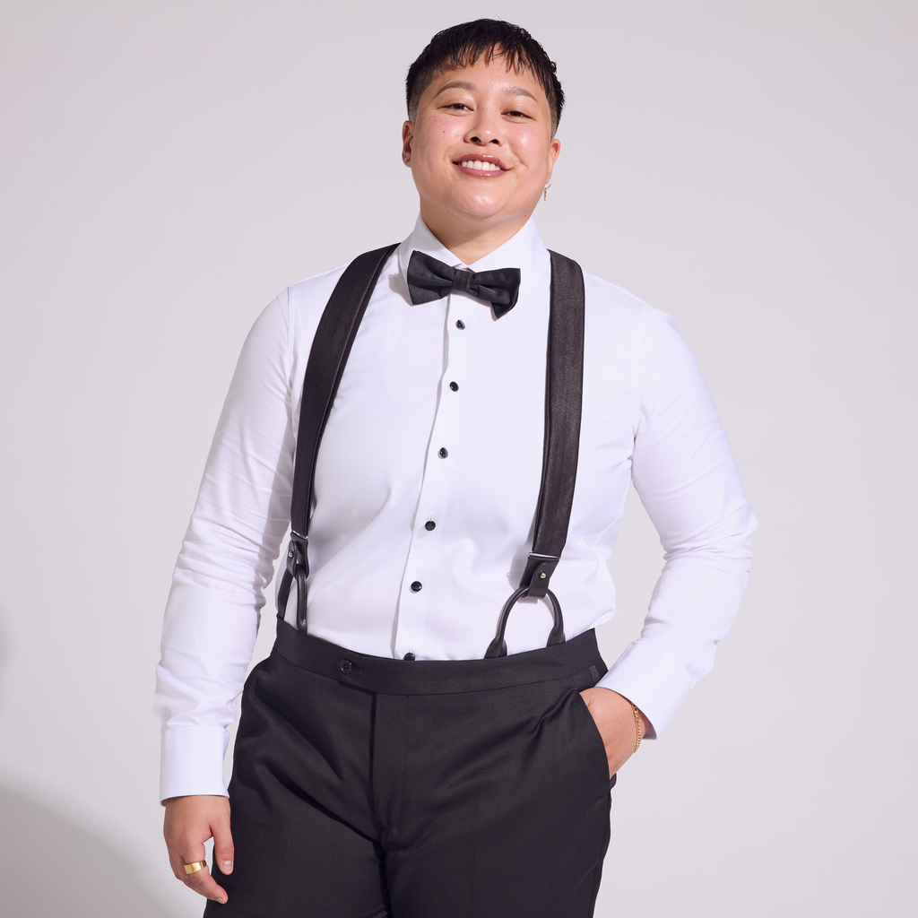A person wearing a white shirt and black tuxedo pants with black satin suspenders and a bow tie.