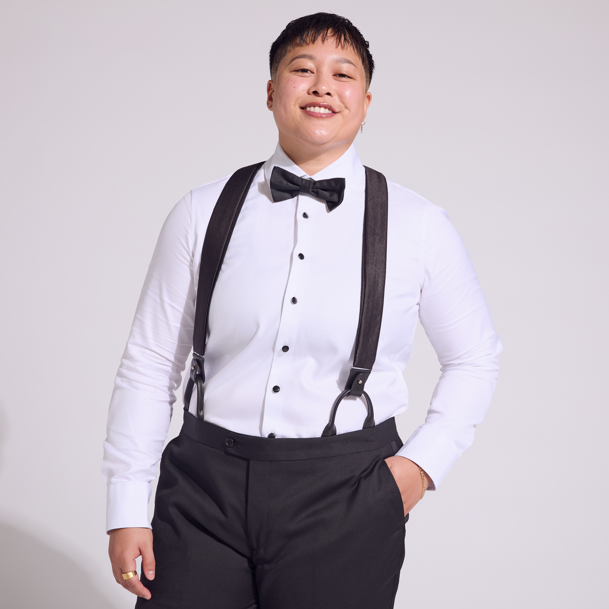 A person wearing a white shirt and black tuxedo pants with black satin suspenders and a bow tie.