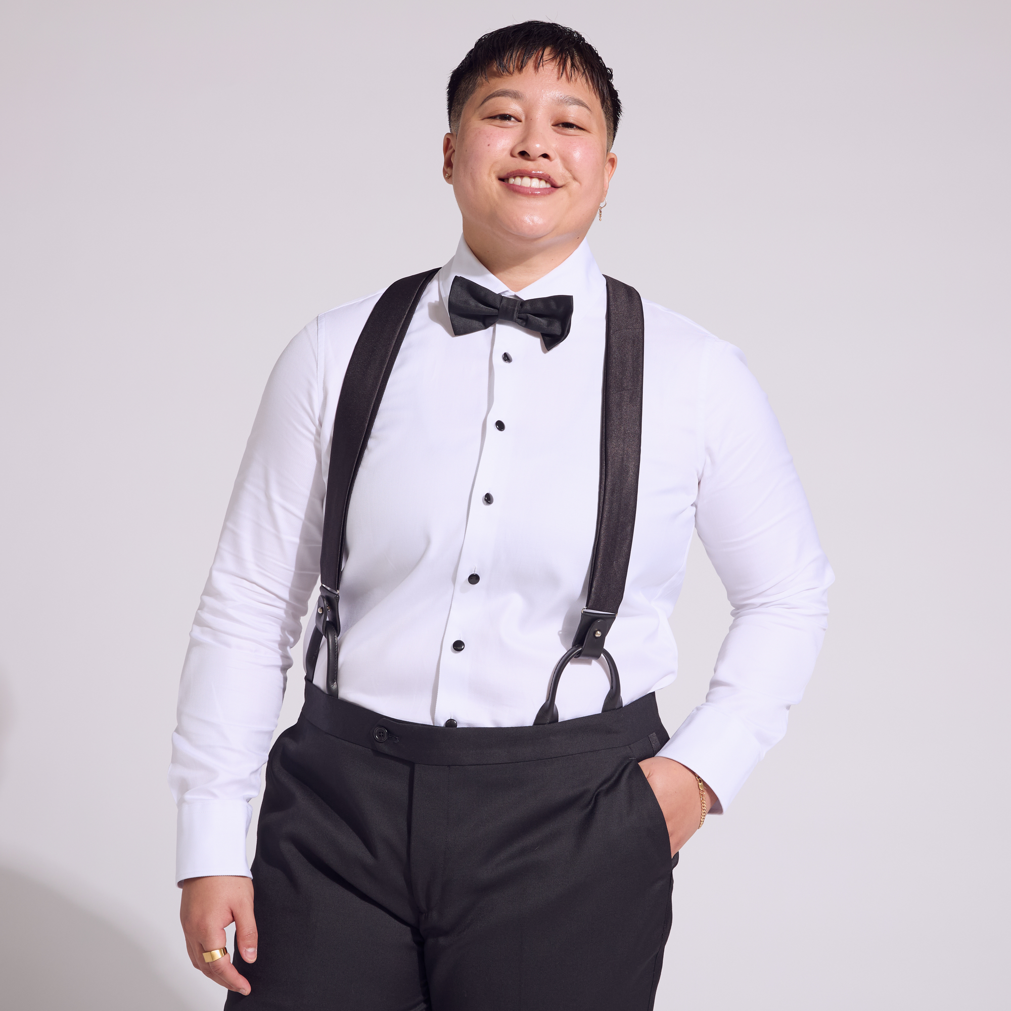 A person wearing a white shirt and black tuxedo pants with black satin suspenders and a bow tie.