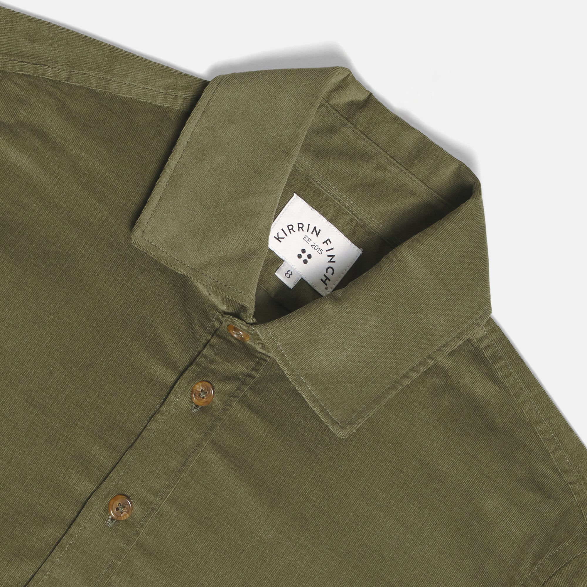 Close-up of an olive green button-down shirt collar with a visible Kirrin Finch brand label, showing the fabric texture and brown buttons on a light gray background. Color: Turtle Corduroy