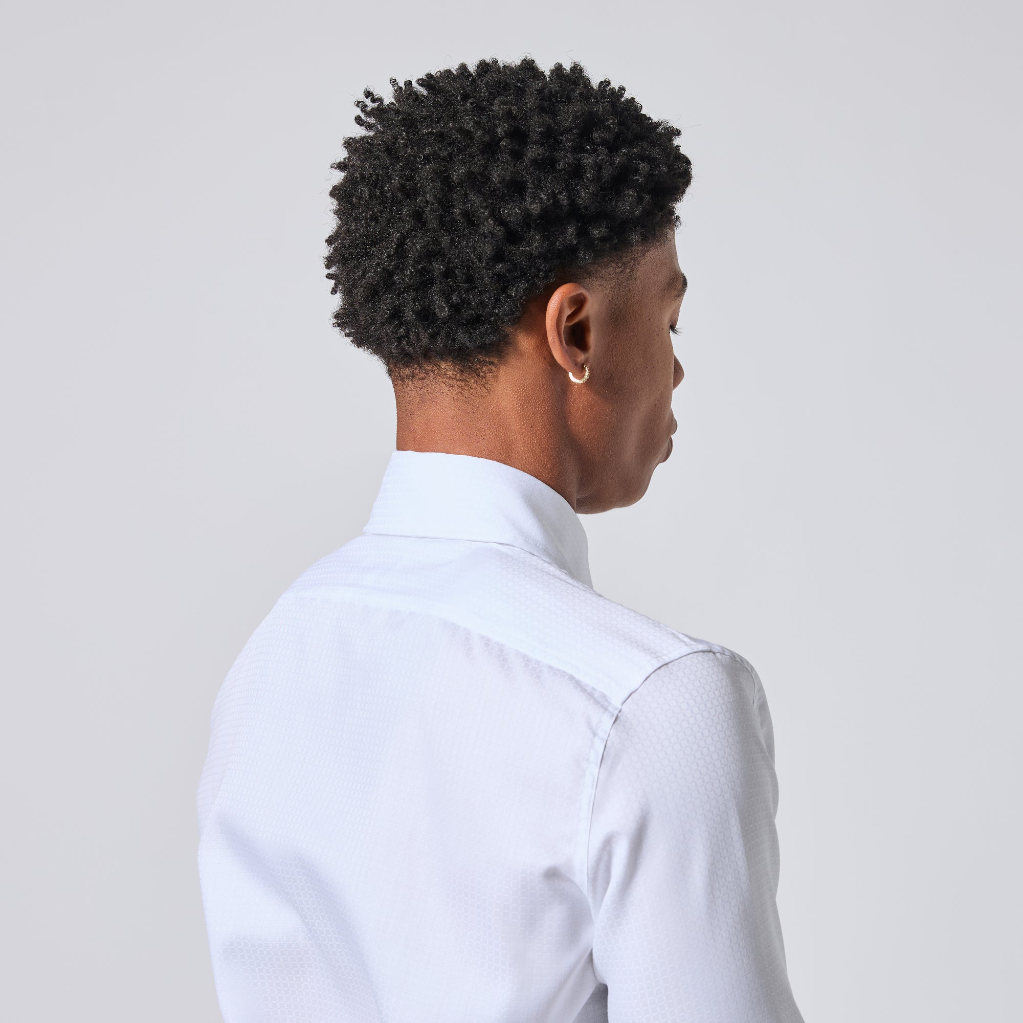 Model shown from the back and side wearing a white textured dress shirt, with short dark curly hair and a small gold hoop earring, against a light gray studio background. Color: White Twill Grid
