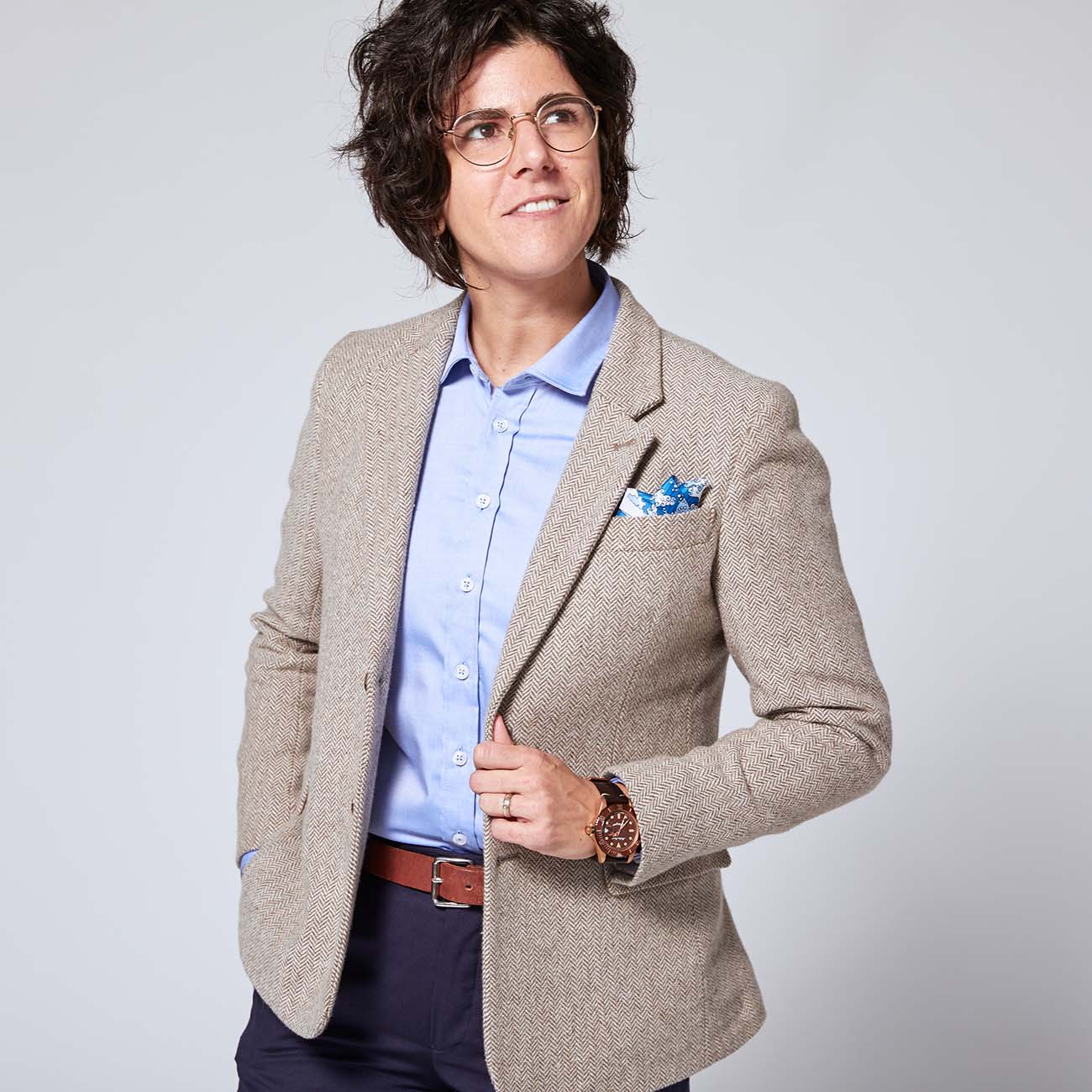 Dapper Androgynous model wearing Taupe Tweed Blazer over a light blue dress shirt and navy dress pants. One hand is in their pants pocket as the other holds the blazer just below the lapel