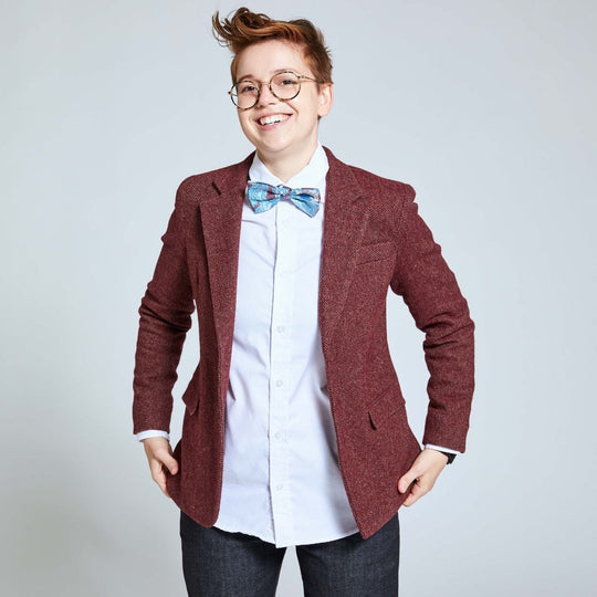 androgynous model wearing burgundy tweed blazer and oceanic bow tie over a white long sleeve dress shirt and blue denim. They are cupping the bottom hem of the blazer.