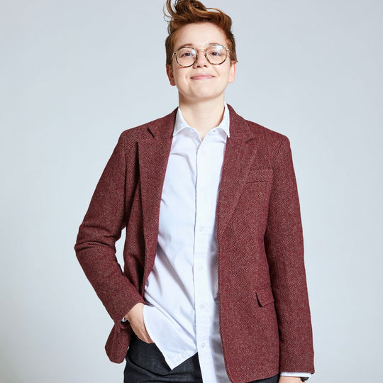 Model wearing Burgundy herringbone tweed blazer with white dress shirt and blue jeans. Their hand is in the left pocket of the denim pants.