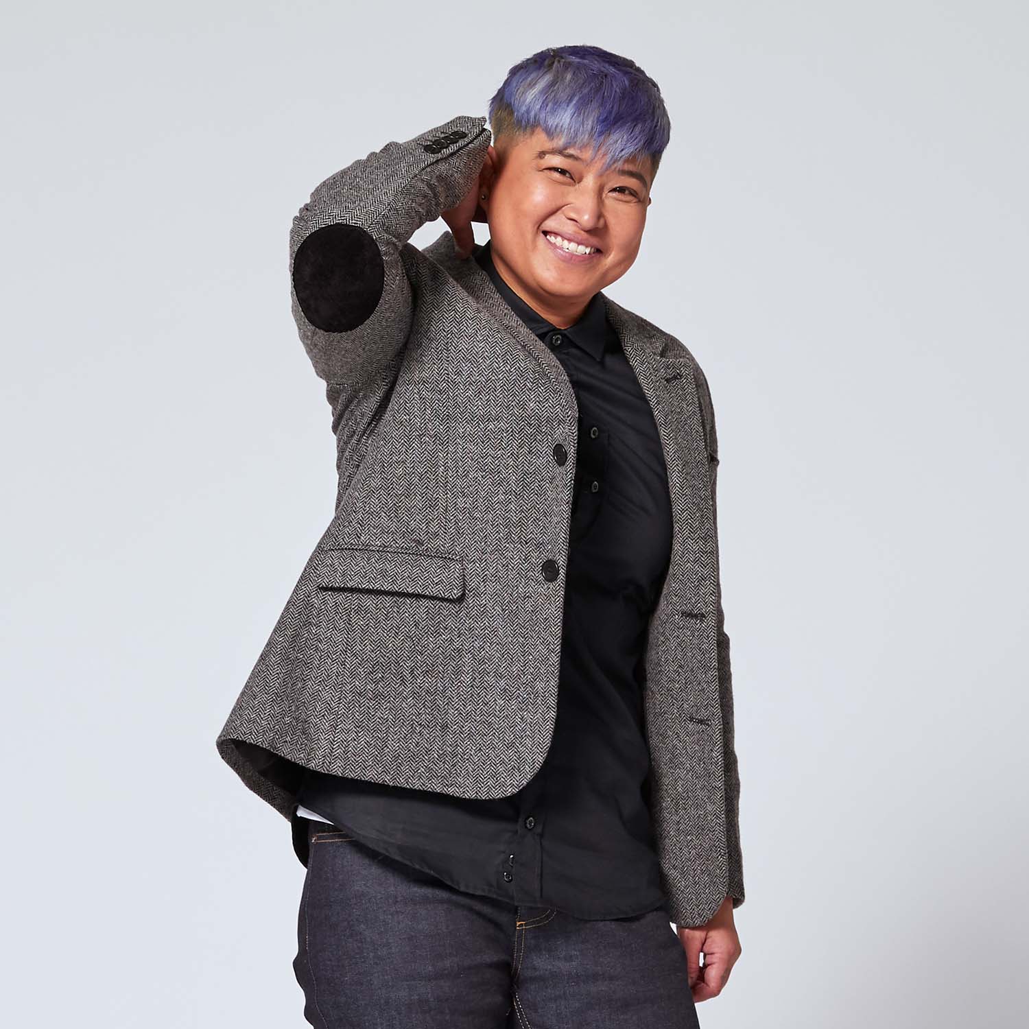 non-binary model wearing the gray tweed blazer and lifting arm to show off the black faux suede elbow patch