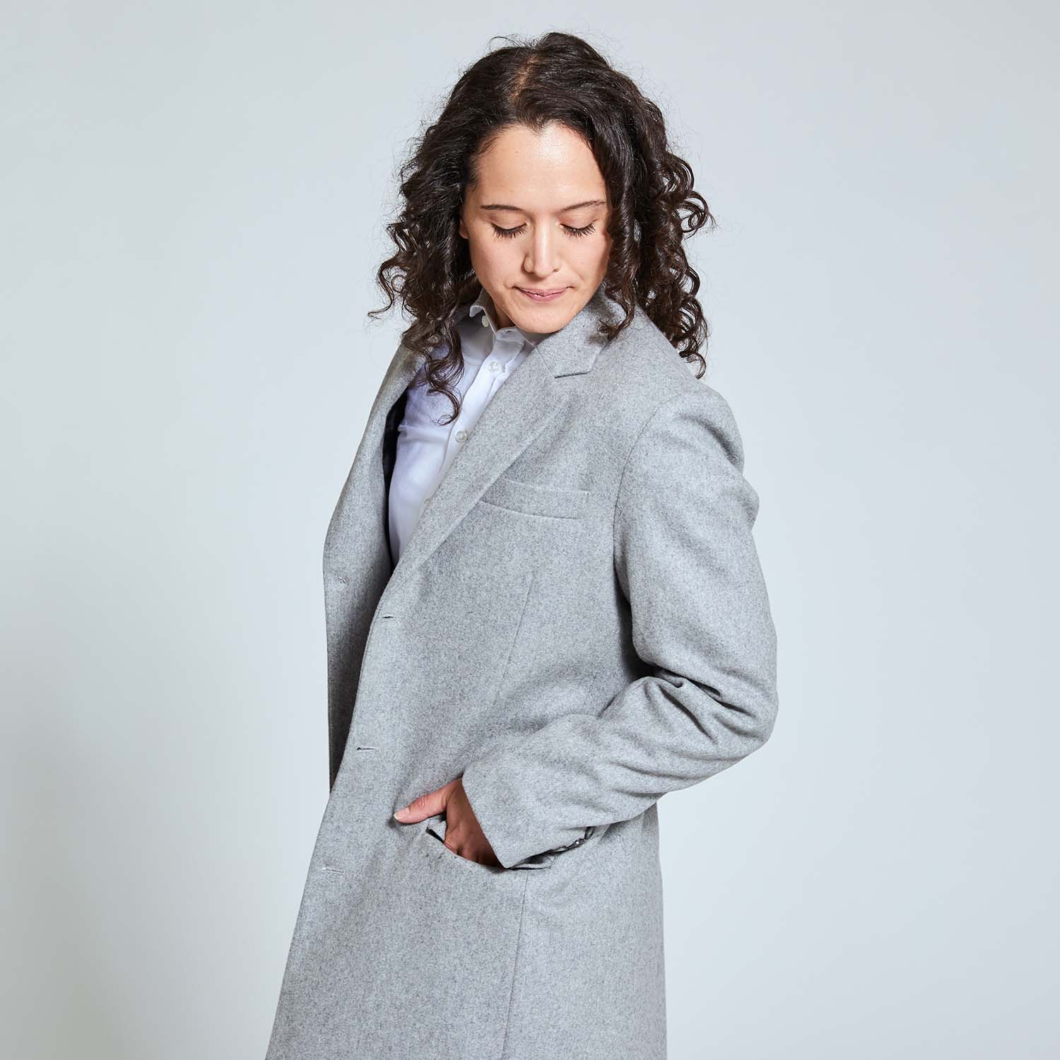 Model with their hand inside gray overcoat pocket while looking down.