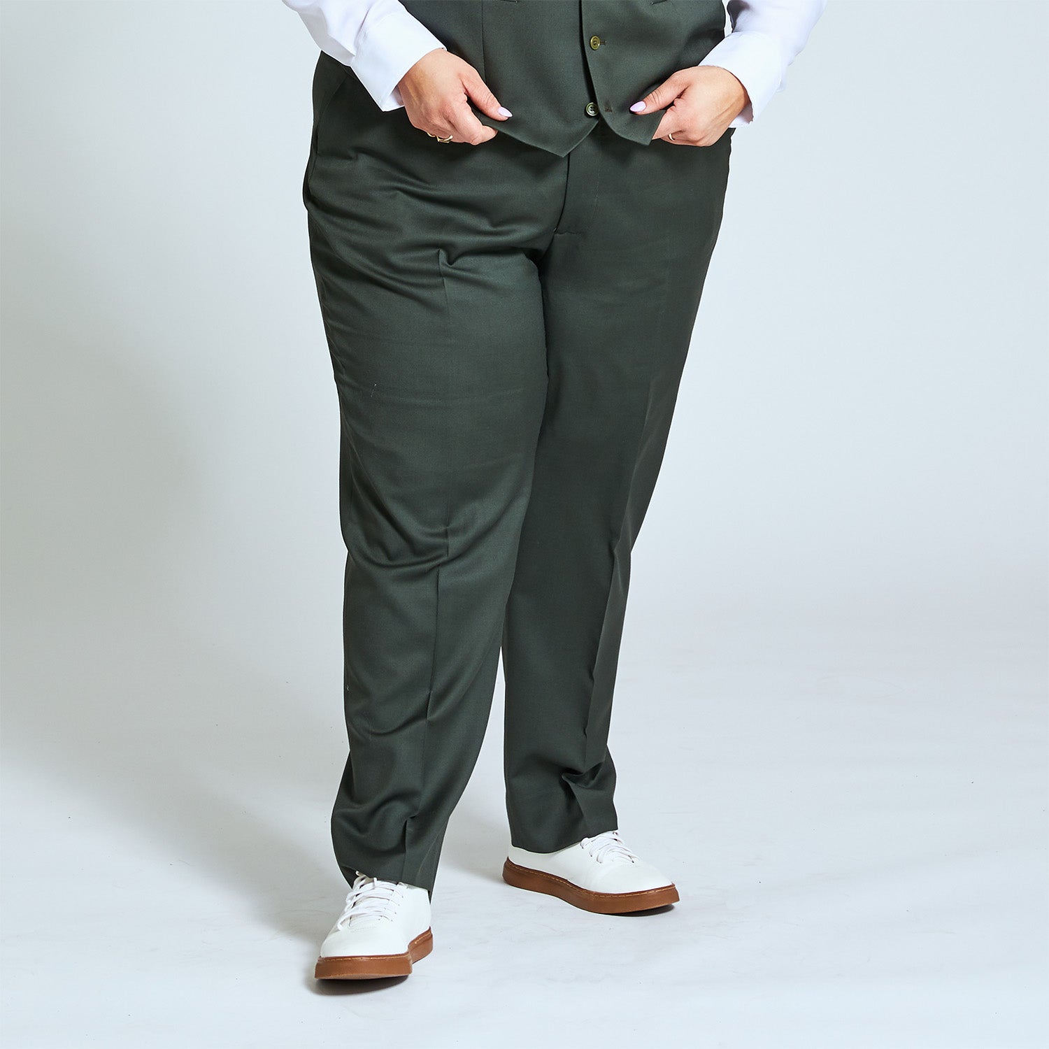 Androgynous model wearing olive dress pants by Kirrin Finch. She is pulling down the matching olive and wearing white sneakers with a gum sole. Neutral studio background.