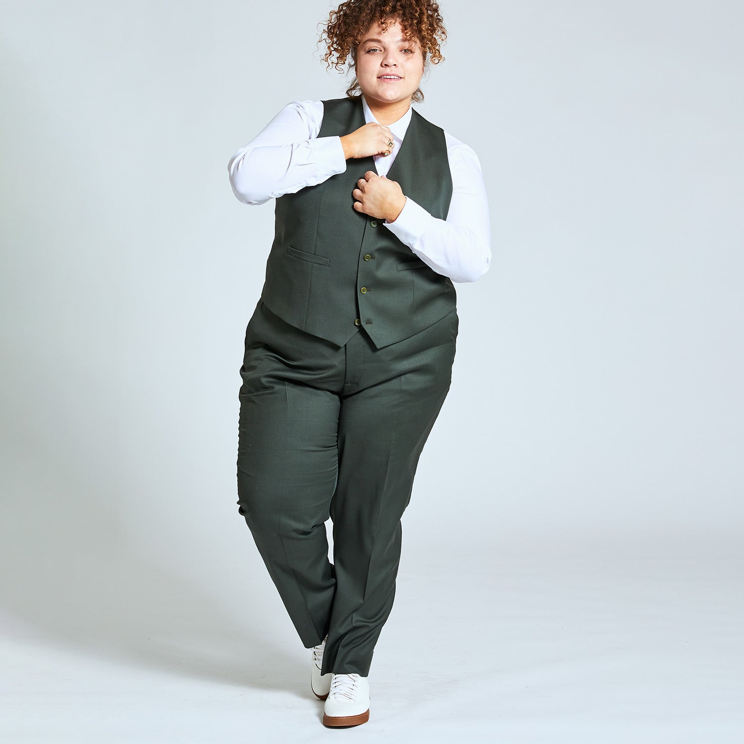 Androgynous model wearing the Georgie olive dress pants and matching olive vest. They are adjusting their white dress shirt buttons and are pictured mid-step. Color: Olive